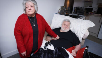 Bu görselde, bir kadın ve bir erkek iç mekânda birlikte poz veriyor. Erkek, yatağında uzanmış durumda ve fiziksel olarak yardıma muhtaç görünüyor. Üzerinde siyah bir tişört var ve yatağında uzanırken televizyon kumandaları gibi eşyalar yanında duruyor. Kadın ise yanında ayakta duruyor, kırmızı bir hırka giymiş ve ifadesi ciddi. Kadının eli, destek verircesine erkeğin göğsüne dokunuyor.