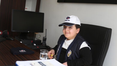 Bu görselde, bir ofis ortamında masa başında oturan küçük bir çocuk yer almakta. Çocuk, polis kıyafeti giymiş ve başında "Polis" yazılı beyaz bir şapka bulunuyor. Masada bilgisayar, telsiz, su bardağı ve bazı belgeler yer almakta. Çocuk gülümsüyor ve elinde bir kalem ya da küçük bir nesne tutuyor.