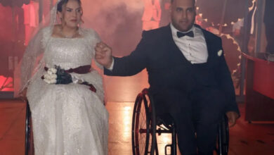 Fotoğrafta, gelinlik giymiş bir kadın ve smokin giymiş bir erkek yan yana tekerlekli sandalyelerinde oturuyor. El ele tutuşarak düğün salonuna giriş yapıyorlar. Gelin, beyaz taş işlemeli bir gelinlik ve taç takarken; damat, siyah smokin ve papyon giymiş. Arka planda sis efekti ve loş ışıklar bulunuyor, bu da sahneye dramatik ve özel bir hava katıyor. Fotoğraf, özel bir an olan düğün töreninden alınmış gibi görünüyor ve çiftin mutlu bir birlikteliğe adım attığını yansıtıyor.
