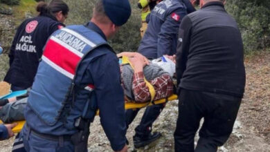 Görselde, zorlu bir arazi ortamında gerçekleşen kurtarma operasyonu görülüyor. Jandarma, sağlık ve kurtarma ekipleri, sedye üzerinde yaralı bir kişiyi taşımakta. Yaralının başında bir boyunluk bulunuyor ve üstü kazak ve kareli gömlek ile örtülü. Arka planda ise yeşillik ve engebeli bir arazi mevcut. Olayın ciddi bir kurtarma operasyonu olduğu anlaşılıyor.