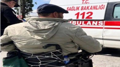 Bu görselde, tekerlekli sandalyede oturan bir kişi sırtı dönük şekilde görüntülenmiş. Kişi açık renk bir mont ve şapka giymekte. Hemen önünde, üzerinde “T.C. SAĞLIK BAKANLIĞI 112 AMBULANS” yazılı bir ambulans aracı bulunuyor. Arka planda bir başka kişi daha yer almakta. Görüntü, acil bir durum ya da yardım çağrısı bağlamında çekilmiş izlenimi veriyor.