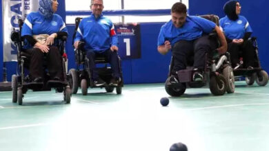 Bu görselde, tekerlekli sandalyede bulunan dört sporcu bir spor salonunda boccia oyunu oynarken görülüyor. Sporcular mavi formalar giymiş ve odaklanmış şekilde oyuna katılıyorlar. Ortamda “KOCAELİ” yazılı bir pankart dikkat çekiyor. Zeminde boccia topları yer alıyor ve bir oyuncu topu ileri doğru atmak üzere.