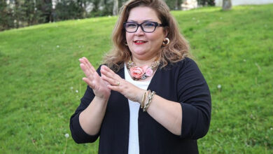 Bu fotoğrafta, yeşillik bir alanda ayakta duran bir kadın yer almakta. Kadın, iki elini işaret diliyle bir şey anlatır şekilde tutmuş. Üzerinde siyah bir ceket ve beyaz bir bluz var; boynunda büyük bir çiçekli kolye dikkat çekiyor. Gözlük takıyor ve hafif gülümseyerek kameraya bakıyor. Kadının bu duruşu ve el hareketleri, işaret diliyle iletişim kurduğunu düşündürüyor.