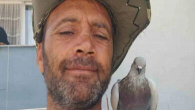 Bu görselde, bir adamın yakın plan çekilmiş bir selfie’si yer alıyor. Başında geniş kenarlı bir şapka var ve yüzünde hafif bir gülümseme ifadesi bulunuyor. Adamın omzuna konmuş bir güvercin dikkat çekiyor; kuş oldukça sakin görünüyor. Arka planda bir bina cephesi ve pencere görülüyor.