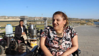 Fotoğrafta ön planda akülü tekerlekli sandalye üzerinde oturan genç bir kadın var. Kadının arkasında ise bir çeşit demirden yapılmış römork ve çay malzemelerinin başında aküle tekerlekli sandalye ile bekleyen bir adam var.