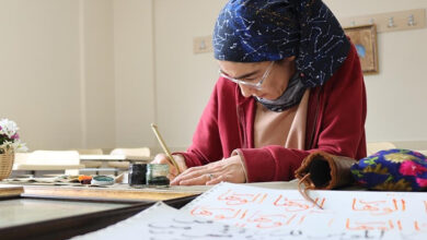 Görselde, başörtülü bir kadın hat sanatıyla uğraşırken görülüyor. Kırmızı bir hırka giymiş olan kadın, bir sınıf ortamında masaya oturmuş ve elinde bir hat kalemiyle dikkatlice yazı yazıyor. Masanın üzerinde mürekkep, kalem ve Arap harfleriyle yazılmış kâğıtlar bulunuyor. Arka planda ise boş sıralar ve duvarda asılı askılıklar mevcut. Kadının sanata olan ilgisi ve çalışmasına olan yoğun dikkati hissediliyor.