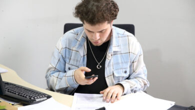 Görselde, genç bir adam bir masa başında oturuyor. Üzerinde mavi tonlarında kareli bir gömlek ve siyah tişört bulunuyor. Boynunda ay yıldız sembollü kolyeler takılı. Masada çok sayıda kâğıt ve evrak yer almakta; adam bir yandan önündeki belgeye dokunuyor, diğer yandan telefonunu tutarak dikkatlice ekrana bakıyor. Yan tarafında bir bilgisayar klavyesi de görülüyor. Ortam bir ofis ya da çalışma alanı izlenimi veriyor.