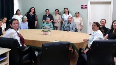 Görselde, büyük bir masa etrafında toplanmış çeşitli yaş gruplarından kadın ve erkekler görülüyor. Bazıları otururken bazıları ayakta duruyor ve kamera karşısında gülümsüyorlar. Masanın üzerinde süs bitkileri yer almakta. Arka planda açık renk duvar, kapı ve üzerinde "ARDUÇ" ve "LADİN" yazılı tabelalar bulunuyor. Bu görüntü, bir bakım merkezi veya sosyal hizmet kuruluşunda gerçekleştirilen bir ziyareti yansıtıyor olabilir.