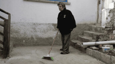 Görselde, dış mekânda bir avluda süpürgeyle temizlik yapan orta yaşlı bir kadın yer alıyor. Kadın, desenli pantolon ve koyu renk bir üst giymiş. Arka planda eski yapılı bir evin merdivenleri, beyaz duvarları ve mavi çerçeveli bir pencere görülüyor. Kadının ifadesi ciddi ve kararlı. Bu kare, günlük yaşam mücadelesini sürdüren, bağımsız ve çalışkan bir bireyi yansıtıyor.