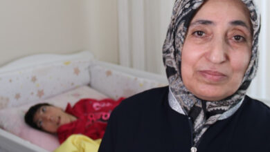 Bu duygusal görselde, ön planda başörtülü, olgun yaşta bir kadın gülümseyerek kameraya bakıyor. Arka planda ise bir çocuk (muhtemelen genç yaşta) özel bir bakım yatağında yatıyor; çocuğun engelli olduğu görülmekte. Kadının bakışlarındaki yorgunlukla karışık sevgi ve azim, yaşam mücadelesini yansıtıyor.