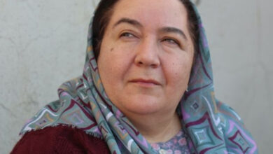 Bu görselde, başörtülü bir kadın yüzünde düşünceli bir ifade ile hafifçe yukarıya doğru bakıyor. Üzerinde desenli, açık renkli bir başörtüsü ve bordo bir giysi var. Arka plan sade, açık renkli bir duvar ile sınırlı. Kadının yüzünde huzurlu ve vakur bir duruş göze çarpıyor.