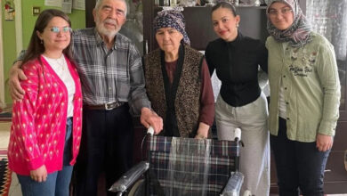 Görselde, bir ev ortamında beş kişi kameraya poz veriyor. Ortada yaşlı bir kadın ve yaşlı bir adam yer alıyor; kadın başörtüsü takıyor ve adamın koluna tutunmuş. Yaşlı çiftin yanında üç genç kadın bulunuyor; biri pembe hırka giymiş, biri siyah üst ve gri eşofman giymiş, diğeri ise başörtülü ve yeşil gömlekli. Önde bir tekerlekli sandalye görülüyor. Görsel, kuşaklar arası dayanışmayı, ziyaret ve destek temalı sıcak bir anı yansıtıyor.