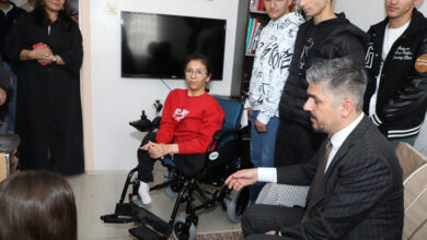 Görselde, kırmızı bir kazak giymiş genç bir kadın tekerlekli sandalyede oturuyor. Etrafında takım elbise giymiş bir adam ve birkaç genç erkek bulunuyor. Oda ev ortamında gibi görünüyor; duvarda bir televizyon, kitaplık ve halı mevcut. Arkada birkaç kişi daha izleyici konumunda duruyor. Görsel, sosyal bir ziyaret veya anlamlı bir yardım etkinliği sırasında çekilmiş izlenimi veriyor.