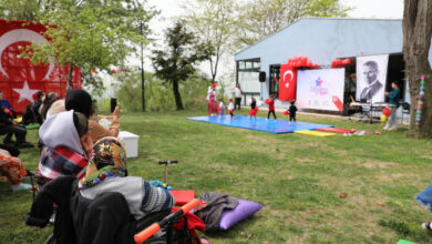 Fotoğrafta, açık havada düzenlenen bir etkinlik görülüyor. Çim alanda çocuklar sahnede gösteri yaparken, izleyiciler arasında tekerlekli sandalyede oturan bireyler de bulunuyor. Sahnenin arkasında Türk bayrağı, Atatürk posteri ve “ÖZGEM” yazılı bir afiş yer alıyor. Etkinlik alanı balonlar ve süslemelerle donatılmış. Bahar havası hâkim, ağaçlar yeşermiş. Aileler çocukları izlerken fotoğraf çekiyor, samimi ve neşeli bir atmosfer var.