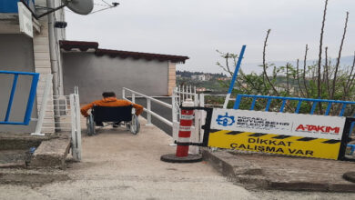 Görselde, turuncu mont giymiş bir kişi tekerlekli sandalyeyle bir rampada ilerliyor. Rampanın iki yanında beyaz metal korkuluklar bulunuyor. Sol tarafta bir bina, çanak anten ve mavi korkuluklar görülüyor. Sağ tarafta ise "Dikkat Çalışma Var" yazılı bir uyarı tabelası ve "Kocaeli Büyükşehir Belediyesi" logosu yer alıyor. Arka planda ağaçlar ve açık hava manzarası bulunuyor. Görüntü, engelliler için yapılan altyapı çalışmalarıyla ilgili olabilir.