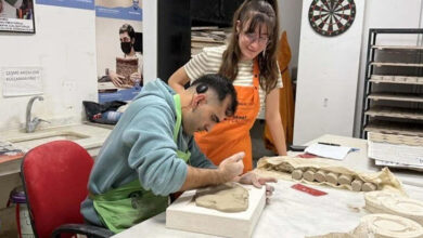 Bu fotoğrafta, bir seramik atölyesinde çalışan bir birey ve ona yardımcı olan bir eğitmen yer alıyor. Oturan kişi, kil ya da seramik bir malzeme üzerine detaylı bir çalışma yaparken, yanında duran kişi ona destek oluyor ve çalışmasını izliyor. Arka planda çeşitli seramik işleri, aletler ve duyuru panoları dikkat çekiyor. Bu görsel, engelli bireylerin sanat yoluyla kendilerini ifade edebilmeleri ve destekleyici bir ortamda yeteneklerini geliştirebilmelerinin güzel bir örneğini yansıtıyor.