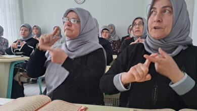 Bu görselde, başörtülü kadınlardan oluşan bir grup bir sınıf ortamında oturuyor. Ön sıradaki iki kadın, elleriyle işaret dili kullanarak iletişim kuruyor. Masalarının üzerinde açık Kur'an-ı Kerimler bulunuyor. Arka planda da benzer şekilde Kur’an-ı Kerim okuyan ya da işaret diliyle öğrenmeye katılan diğer kadınlar yer almakta. Görseldeki atmosfer, dini eğitim alan işitme engellilere yönelik bir kursu yansıtıyor.
