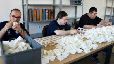 Görselde, bir atölyede masa başında çalışan üç birey yer alıyor. Masanın üzerinde beyaz plastik parçalar ve kutular bulunmakta. Çalışan bireyler dikkatlice bu parçaları birleştiriyor ya da ayıklıyor. Raflarda çeşitli kutular ve malzemeler yer almakta. Görsel, engelli bireylerin üretken bir şekilde iş hayatına katılımını ve onların istihdam edildiği destekleyici bir iş ortamını yansıtıyor.