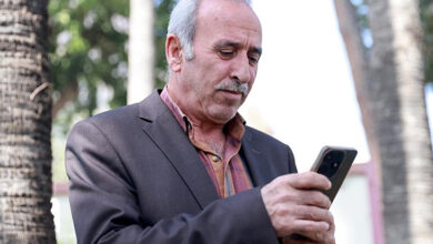 Bu görselde, yaşlı bir adam açık havada, ağaçların arasında dururken cep telefonuna bakıyor. Üzerinde koyu renk bir ceket ve kareli gömlek bulunuyor. Adamın ifadesi ciddi ve dikkatli; sanki telefonda önemli bir şeyle ilgileniyor. Arka planda ise bulanık şekilde palmiyeler ve doğa manzarası görülüyor.