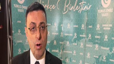 Görselde, takım elbise giymiş bir erkek Yunus Emre Vakfı’nın düzenlediği bir etkinlikte konuşma yaparken ya da röportaj verirken görüntülenmiş. Arka planda, üzerinde “Türkçe Birleştirir” sloganı ve çeşitli şehir isimlerinin yazılı olduğu Yunus Emre Vakfı’na ait yeşil bir tanıtım panosu yer alıyor. Erkek kişi ciddi bir ifadeyle kameraya bakmakta ve üzerinde TBMM rozetine benzer bir pin takılı. Görsel, kültürel veya diplomatik bir etkinlik kapsamında yurt dışında düzenlenen bir tanıtım ya da gösterim sırasında çekilmiş izlenimi veriyor.