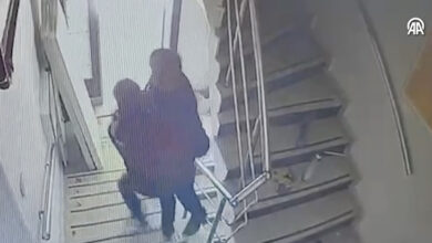 Görsel, bir güvenlik kamerası kaydını andıran, bulanık ve gri tonlarında bir sahneyi göstermektedir. Bir merdiven boşluğunda, iki kişi görülmektedir. Bir kişi, diğerini kollarının arasına almış ve yukarı doğru taşımaktadır. Taşıyan kişi koyu renkli giysiler giymektedir ve taşınan kişinin yüzü tam olarak seçilememektedir.