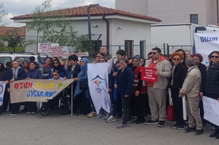 Görselde, çoğunluğu kadınlardan oluşan bir grup insan, kaldırım üzerinde ve bir binanın önünde toplanmış durumda. Ellerinde çeşitli yazılar içeren pankartlar ve bir de üzerinde "Türkiye Özel Sporcular Spor Federasyonu" yazan bir bayrak taşıyorlar. Bazı pankartlarda "Otizm Eylem Planı Uygulansın" ve "Bensiz Kalan Otizm Ortakları" gibi ifadeler okunuyor. Bir kişi tekerlekli sandalyede oturuyor.