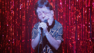 Adam Pearson sahnede mikrofon tutarak şarkı söylüyor.