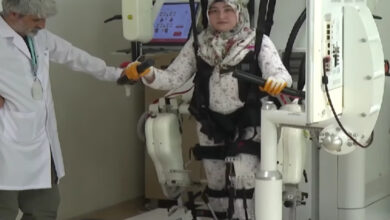 Görselde, bir kadın hasta bir yürüme robotu cihazına bağlı şekilde fizik tedavi görmekte. Cihaz, hastanın bacaklarına destek sağlayarak yürüme egzersizi yapmasına yardımcı oluyor. Kadının elleri tutacaklara bağlı, üzerinde emniyet kemerleri bulunuyor ve bir fizyoterapist ya da sağlık çalışanı da yanında destek veriyor. Arka planda tedavi sürecini izlemeye olanak tanıyan bir monitör yer alıyor.