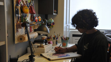 Bu görselde, genç bir kişi ev ortamında bir masada oturmuş resim yapıyor. Kıvırcık saçlı genç, önündeki deftere renkli kalemlerle çizim yaparken odaklanmış bir şekilde çalışıyor. Masanın üzerinde lambalar, kalemlikler, çizim araçları ve düzenli bir çalışma düzeni bulunuyor. Arka duvarda peluş figürler ve kişisel süslemeler yer almakta.