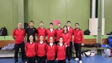 Bu görselde, bir spor salonunda çekilmiş bir grup fotoğrafı yer alıyor. Fotoğrafta, kırmızı eşofman üstleri giymiş toplam 12 kişi bulunuyor. Arka sırada 7 kişi ayakta dururken, ön sırada 5 kadın sporcu diz çökerek poz veriyor. Grup, Türkiye’yi temsil eden bir spor takımı gibi görünüyor; çünkü arka duvarda Türk bayrağı asılı. Fotoğrafın çekildiği yer muhtemelen bir badminton salonu, zira çevrede spor çantaları ve ekipmanlar yer almakta.