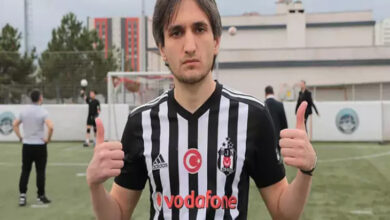 Bu görselde, bir futbol sahasında Beşiktaş forması giymiş genç bir erkek yer alıyor. Oyuncu, her iki başparmağını yukarı kaldırarak poz vermiş. Üzerindeki forma siyah-beyaz çizgili ve göğsünde Türk bayrağı ile Beşiktaş’ın logosu yer almakta. Arka planda futbol oynayan başka kişiler ve bir kale görülüyor.