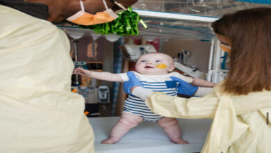 Bu fotoğrafta, hastane ortamında iki sağlık çalışanı, tedavi görmekte olan bir bebeği destekleyerek ayakta tutuyor. Bebek mutlu görünüyor, burnunda medikal bant ve beslenme tüpü var.