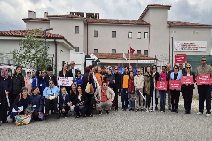 Geniş bir grup insan, büyük, açık renkli bir binanın önünde toplanmış fotoğrafın konusunu oluşturuyor. Bina, üzerinde "T.C. ARİFİYE KAYMAKAMLIGI... (birkaç kelime okunamıyor)... OTİZM MERKEZİ" yazan bir tabelaya sahip. Ayrıca binada Türk bayrağı da asılı duruyor.