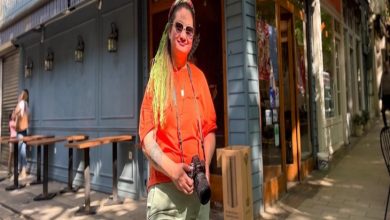 Görselde, bir kadın şehir sokaklarında poz veriyor. Üzerinde turuncu tişört, açık renk pantolon ve boynunda bir fotoğraf makinesi var. Uzun yeşil saçları dikkat çekici ve güneş gözlüğü takıyor. Arka planda kafe ya da restoranlar bulunuyor. Kadın özgüvenli bir duruşla objektife bakıyor.