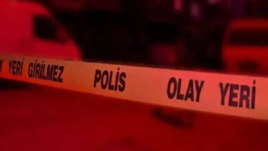 Bu görselde, sarı renkli “POLİS OLAY YERİ – GİRİLMEZ” yazılı emniyet şeridi yer alıyor. Şerit, bir olay yerinin çevresini güvenlik amacıyla kapatmak için kullanılıyor. Arka plan kırmızı ağırlıklı ışıkla aydınlatılmış, muhtemelen gece çekilmiş bir sahne; arkada silüet hâlinde araçlar ve belirsiz yapılar görünüyor.