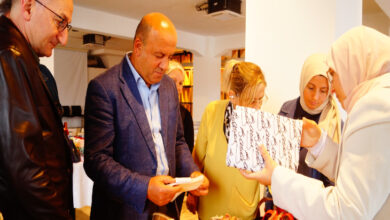 Bu görselde, bir grup insan kapalı bir mekânda düzenlenen el sanatları sergisinde ürünleri inceliyor. Ön planda, farklı yaşlardan erkek ve kadınlar masadaki el emeği ürünlere yoğun ilgi gösteriyor. Sergilenen ürünler arasında el işi çantalar, örgüler ve dekoratif eşyalar yer alıyor. Kadınlardan biri, üzerinde yazılar bulunan bir kumaşı ya da çantayı gösteriyor.