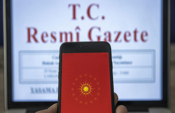 Görselde, ön planda bir cep telefonu ekranı yer almakta. Ekranda kırmızı zemin üzerinde Türkiye Cumhurbaşkanlığı forsunun simgesi görülüyor. Arka planda ise bir dizüstü bilgisayar ekranında “T.C. Resmî Gazete” başlığı dikkat çekiyor. Görsel, Türkiye’de alınan resmî kararların ve duyuruların yayımlandığı Resmî Gazete’ye dijital erişimi temsil ediyor.
