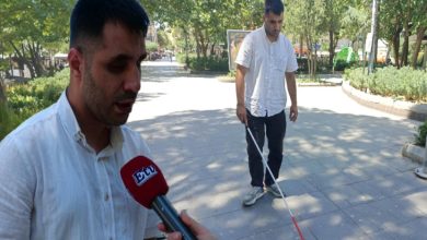 Görselde, açık havada ağaçlarla çevrili bir alanda iki erkek yer alıyor. Önde bulunan kişi mikrofon tutularak röportaj yapıyor; mikrofonun üzerinde "ETC" yazısı görülüyor. Arkada yer alan kişi ise beyaz baston kullanarak yürüyen doğuştan görme engelli bir birey. Her iki kişi de beyaz gömlek giymiş. Görsel, 6 Şubat depremi sonrası Manisa’ya taşınan ve iş arayan görme engelli Kamil Çelik’in kamuoyuna seslendiği bir anı yansıtıyor.