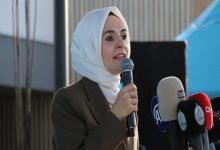 Görselde, başörtülü bir kadın açık havada mikrofonla konuşma yaparken görülüyor. Üzerinde koyu renk bir ceket var. Önünde çeşitli basın kuruluşlarına ait mikrofonlar yer alıyor (TRT, A Haber, DHA gibi). Arka planda mavi bir çadır ve modern bir bina detayı seçiliyor. Kadın ciddi ve etkileyici bir ifadeyle hitap ediyor.