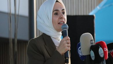 Görselde, başörtülü bir kadın açık havada mikrofonla konuşma yaparken görülüyor. Üzerinde koyu renk bir ceket var. Önünde çeşitli basın kuruluşlarına ait mikrofonlar yer alıyor (TRT, A Haber, DHA gibi). Arka planda mavi bir çadır ve modern bir bina detayı seçiliyor. Kadın ciddi ve etkileyici bir ifadeyle hitap ediyor.