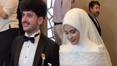 Görselde, gelinlik ve damatlık giymiş bir çift, mutlu bir şekilde yan yana duruyor. Damat papyonlu siyah bir takım elbise giymiş, gülümsüyor. Gelin ise beyaz başörtüsü ve ışıltılı bir gelinlik içinde, hafifçe başını eğmiş. Arka planda birkaç kişi daha görünüyor. Bu kare, bir düğün töreninden, sevgi ve mutluluğun paylaşıldığı özel bir anı yansıtıyor. Damat, aynı zamanda ampute futbolcu olarak biliniyor.