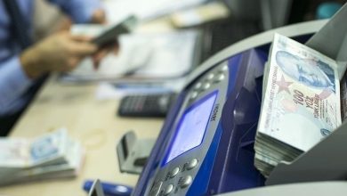 Görselde, bir para sayma makinesi ve yanında yüklü miktarda Türk lirası dikkat çekiyor. Arka planda ise masada oturan bir kişi para sayarken flu şekilde görülüyor. Masanın üzerinde hesap makinesi ve belgeler yer almakta. Görsel, resmi veya kurumsal bir işlem, ödeme ya da mali destek sürecini yansıtan bir sahne sunuyor.