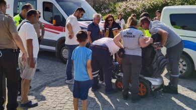 Fotoğrafta akülü tekerlekli sandalye üzerinde oturan engelli bireyin çevresinde sağlık personelleri ve bölgede bulunan insanlar yer alıyor. Görselde engelli birey ambulans ile hastaneye götürülmek için sedyeye taşınıyor.