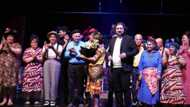 Görselde, sahnede yan yana dizilmiş, çoğunluğu rengârenk ve kostümlü giysiler giymiş bireylerden oluşan bir grup yer alıyor. Ortada bir kadın çiçek buketi tutuyor, yanında ise takım elbiseli bir adam duruyor. Oyuncular, engelli bireylerden oluşuyor ve seyircilerden büyük alkış almışa benziyorlar. Bu sahne, bir tiyatro oyununun sonunda alkışlarla karşılanan, emek ve sanatla dolu bir başarıyı temsil ediyor.