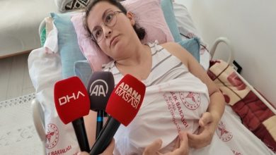 Görselde, hastane yatağında yatan genç bir kadın yer alıyor. Üzerinde Sağlık Bakanlığı’na ait hastane örtüsü var, yüzünde yorgun ama dirayetli bir ifade bulunuyor. Gözlük takıyor ve açık renkli bir atlet giymiş. Kadının önünde mikrofonlar tutulmuş – DHA, AA ve Adana Mavisi haber ajanslarına ait mikrofonlar net şekilde görülüyor.