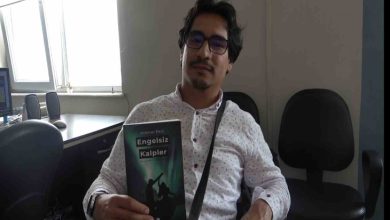 Görselde, gözlüklü bir erkek birey iç mekânda bir sandalyede oturuyor ve elinde “Engelsiz Kalpler” adlı kitabı tutuyor. Üzerinde beyaz, desenli bir gömlek var ve çapraz askılı bir çanta takıyor. Arka planda ofis ortamını andıran masa, bilgisayar ve boş sandalyeler görülüyor. Kitabın kapağında engelli bireylerin silüetleri yer almakta.