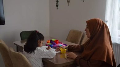 Fotoğrafta, orta yaşlarda olan bir kadın ve bir kız çocuğu masanın etrafında oturuyor. Çocuk önündeki kitaba boyama yapıyor ve masanın üzerinde farklı renklerde kutular var.