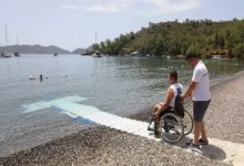 Görselde, bir engelli birey tekerlekli sandalyesiyle denize uzanan özel bir rampada, yanında ona eşlik eden bir kişiyle birlikte görülüyor. Arka planda sakin bir koy, yüzen insanlar ve demirlemiş tekneler var. Bu kare, Muğla’daki “Engelsiz Plajlar” projesi kapsamında engelli bireylerin denize erişimini kolaylaştırmak için yapılan düzenlemeleri yansıtıyor.