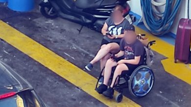 Görselde, bir feribotun araç garajında yer alan iki kişi görülüyor. Biri tekerlekli sandalyede oturan bir çocuk, diğeri ise arkasında oturan ve onu kucaklayan yetişkin bir kadın muhtemelen annesi. İkisi de yazlık giysiler içinde ve telefonlarına bakıyorlar. Etraflarında motosiklet, hortum, bavul ve mavi plastik bidonlar var. Ortam, sıcak ve havasız bir garaj katını andırıyor, zemindeki sarı çizgiler ve kirli beton yüzey dikkat çekiyor.