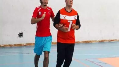 Görselde bir spor salonunda iki genç erkek yan yana koşuyor. Sol taraftaki kişi mavi şort ve kırmızı tişört giymiş; sağ taraftaki ise turuncu-siyah forma giyiyor ve üzerinde "7" numarası yer alıyor. Sağdaki bireyin gözleri kapalı görme engelli olduğu anlaşılıyor; diğer kişi ona rehberlik ediyor. İkili, antrenman ya da sportif bir etkinlik sırasında uyumlu şekilde hareket ediyor. Arka plan sade bir duvar ve spor salonu zeminiyle çevrili.